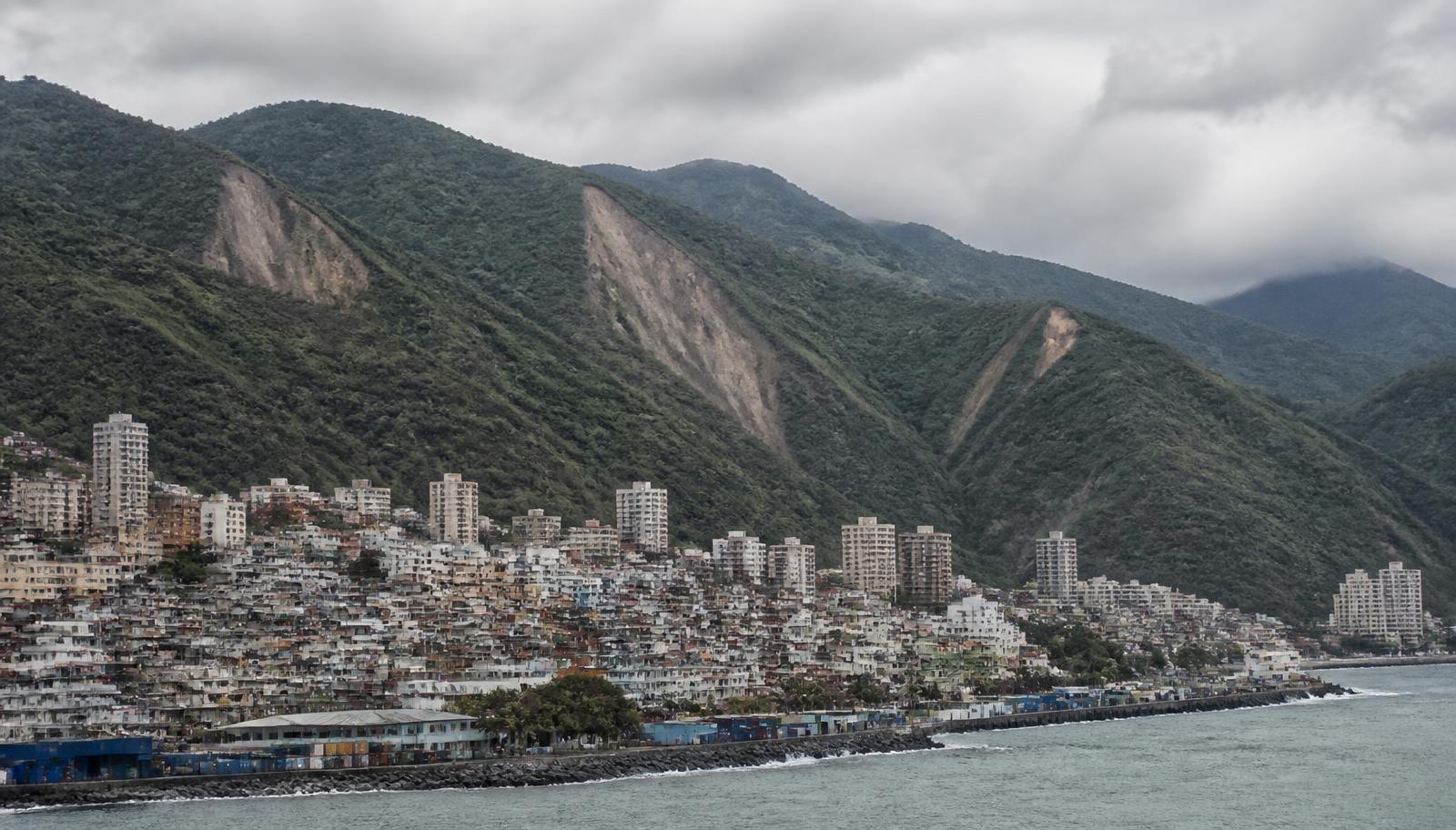 Ciudad costera de La Guaira comprimida entre el mar Caribe y las laderas abruptas de la Cordillera de la Costa, con cicatrices de deslizamiento en las pendientes.