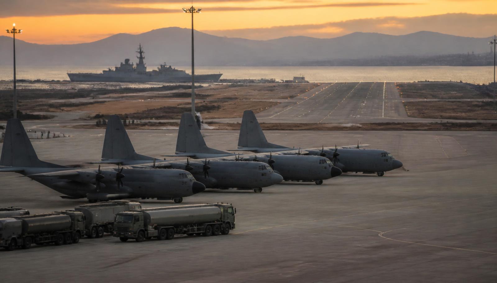 Plataforma de una base militar del sur de España al atardecer, con aviones de transporte estacionados, camiones cisterna y un buque de guerra al fondo.