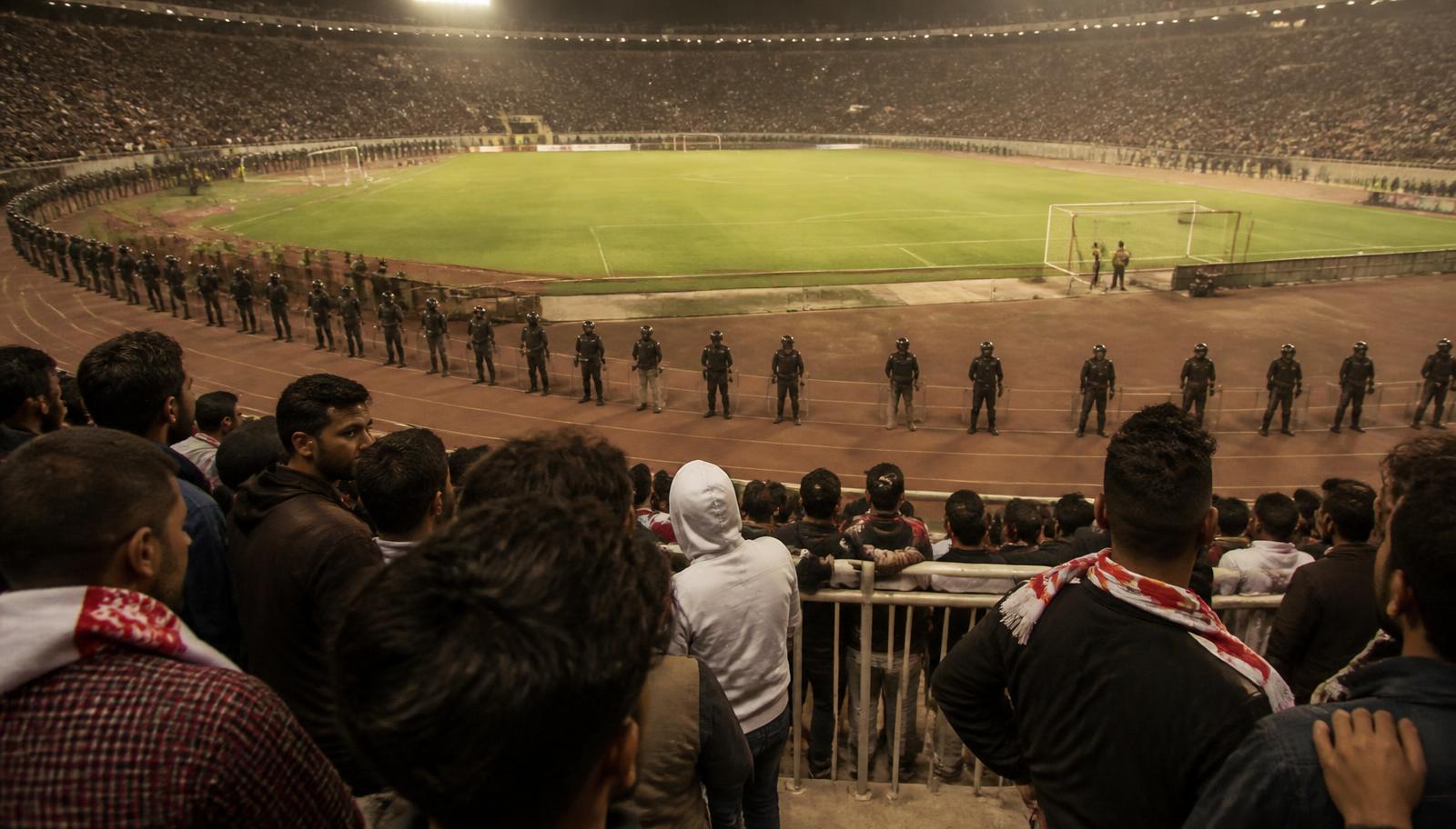 Estadio norteafricano lleno durante un torneo continental, con un cordón de antidisturbios desplegado en la pista de atletismo frente a la grada y el césped vacío.