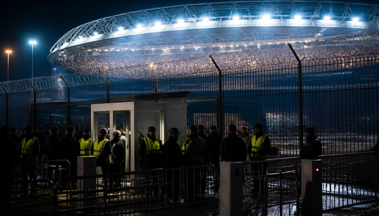 Estadio moderno iluminado de noche visto desde el exterior tras un perímetro de seguridad con vallas, control de acceso y personal de seguridad.